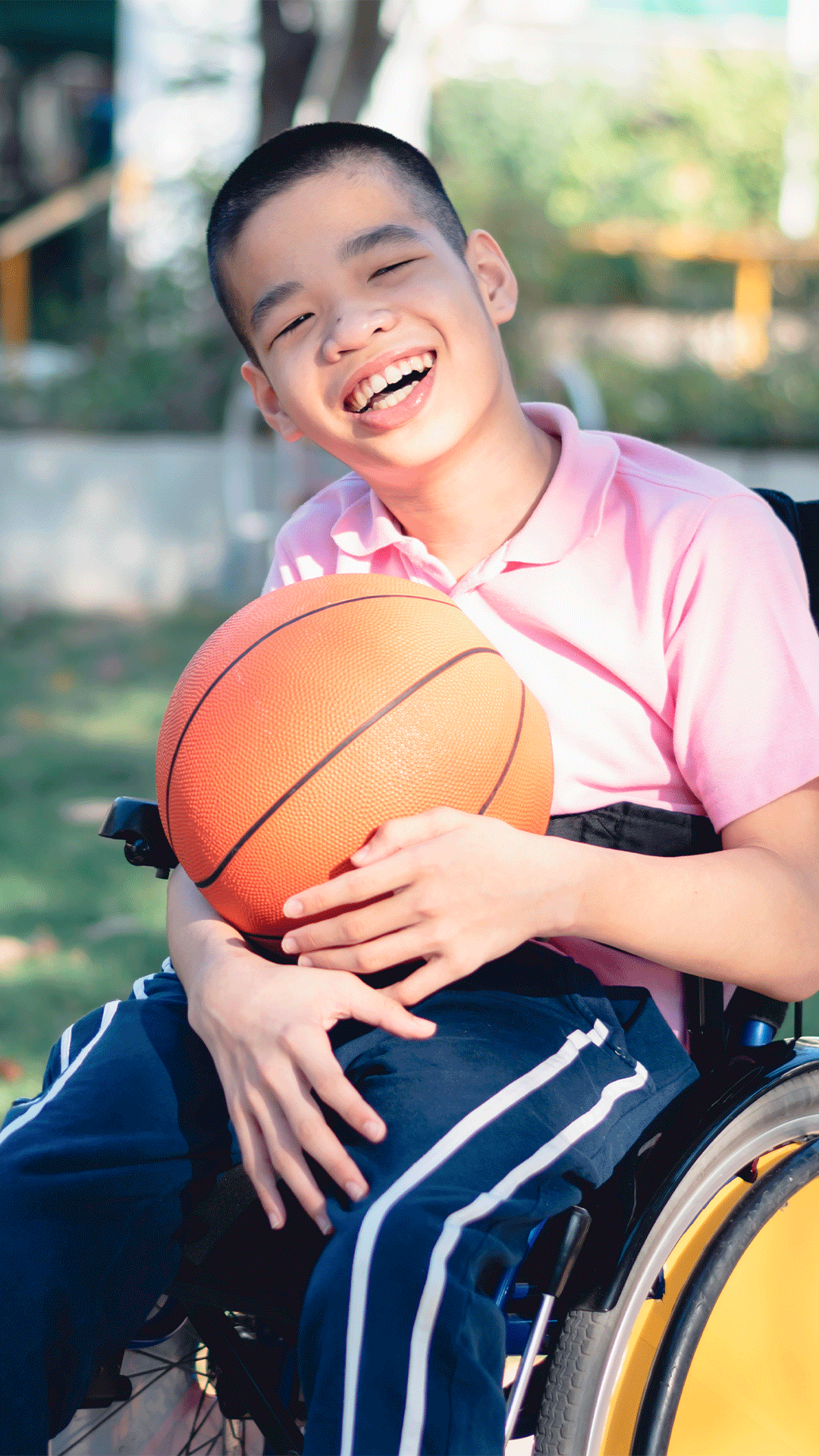 Smiling child using a wheelchair while holding a basketball outdoors, representing inclusive disability support and community participation services.